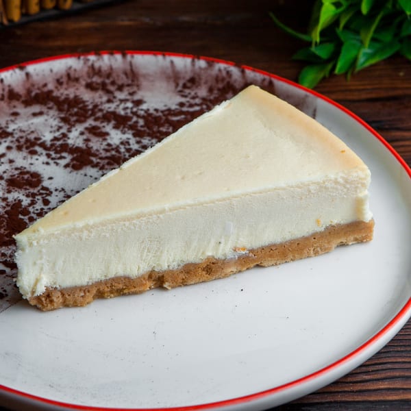 Slice of classic cheesecake with a graham cracker crust and cocoa powder dusting on a white plate.
